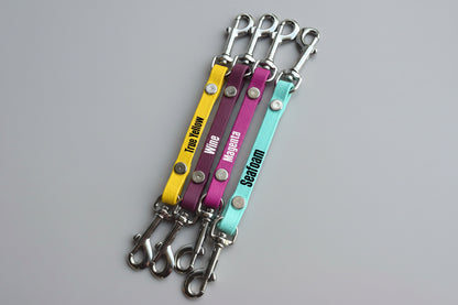 Four colorful keychains with metal hooks on a wooden surface, labeled 'True Yellow', 'Purple', 'Magenta', and 'Seagreen'.